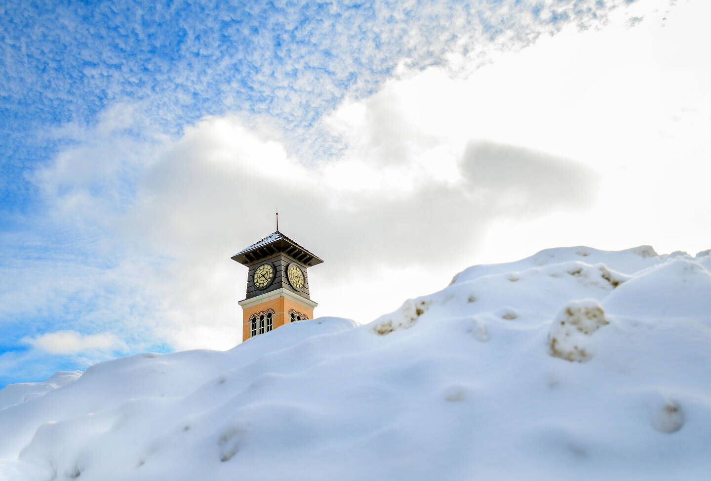 A clock tower rises above a snow-covered hill under a bright, partly cloudy sky.